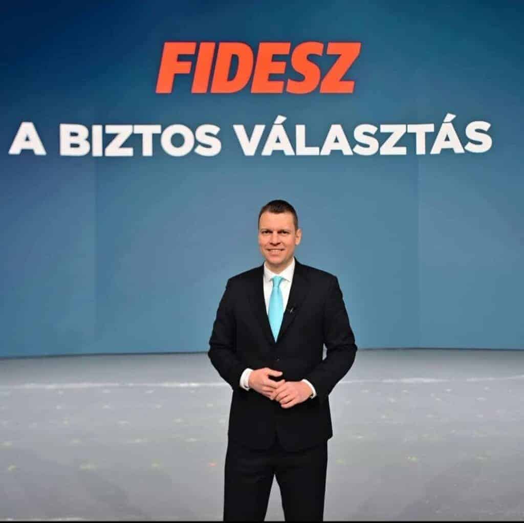 Az a képen egy férfi áll, fekete öltönyben, fehér ingben és kék nyakkendőben, háttérben a FIDESZ-es kampány szlogennel.