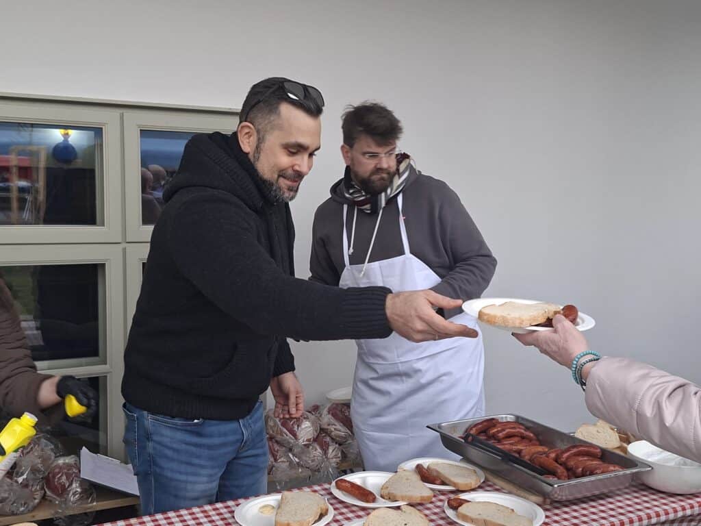 Egy férfi kap egy szelet kolbászt egy fesztiválon, középen egy másik férfi segít.